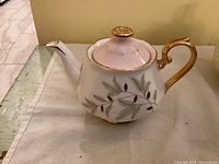 Front side view of the small white Royal Albert Braemar teapot showing gold-accented handle and spout with glued broken tip, leaf and berry pattern on body.