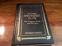 Front cover of The Ultimate Hitchhiker’s Guide hardcover book by Douglas Adams with gold embossed title on black leather-textured cover.