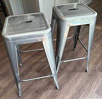 Pair of two metal grey backless counter stools with square seats and cutout handles, viewed side angle on a wood floor.
