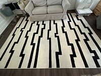 Full view of synthetic fiber area rug with black geometric stepped line design, laid out beneath couches on dark wood floor.