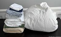 Stack of assorted bedding items including sheets, pillowcases, blankets, a coverlet, and a duvet on a dark surface.