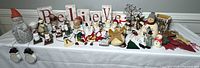 Wide view of multiple Christmas decor items including snowmen figurines, wooden 'Believe' signs, metal tree decorations, and hanging ornaments on a white tablecloth.