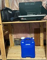 Coleman propane grill/stove on wooden shelf with black Coleman carrying case. Blue Reliance Aqua-tainer water jug below shelf.