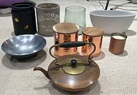 Photo of the miscellaneous decor items showing vintage copper kettle, utensil holder marked 'Parisi Frances 1893', oval white bowl, small candle holder, two hammered copper containers with wooden lids, black pot with bee decorations, green glass, and a patterned shallow metal bowl.