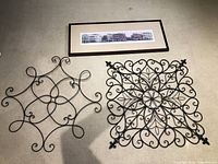 Two metal wall art pieces on beige carpet alongside a panoramic printed artwork in frame with cream matting and brown border