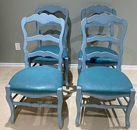 Four blue painted wooden dining chairs placed in a group, showing chairs with teal vinyl seats and scalloped ladder backs.