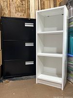 Black 3 drawer metal filing cabinet with silver handles next to a white wooden 3 tier shelf, stored indoors on a concrete floor with plywood wall background.