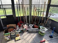 Photo showing comprehensive assortment of gardening tools and supplies grouped indoors near a window. Visible items include shovels, rakes, plant cages, poles, Scotts seed bag, chicken wire roll, and handheld leaf blower.