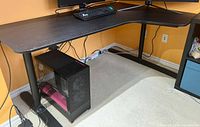 Full view of black L-shape desk with wood top and metal legs, showing overall condition and setup with computer tower below