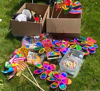 Full view of various colorful windmill spinners, whirly gigs, sidewalk chalk, and Nerf toy displayed on grass with two cardboard boxes containing some items.