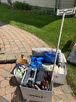 Photo showing boxes containing painting trays, blue tarps, drop cloths, rollers, and other painting supplies on a brick patio.