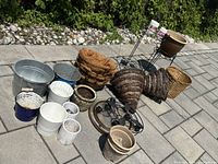 Full lot of assorted plant accessories outdoors on pavement with greenery behind, showing varied planters, pots, hanging baskets, and stands.
