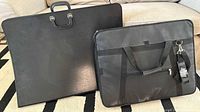 Large black portfolio case and black CHAMPS bag side by side on a carpeted floor, showing overall condition and size difference.