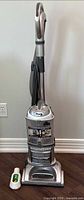 Full view of silver Shark Lift-Away Deluxe upright vacuum cleaner standing against wall, showing its hose and handle with a bottle placed in front for scale.
