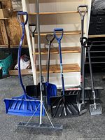Photo of seven assorted shovels and ice tools standing upright on a garage floor with shelving unit behind.