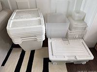 Wide shot showing all storage bins, including four IKEA SORTERA white bins with lids stacked on top of large white under-bed storage bin with wheels, and a grouping of clear plastic bins.