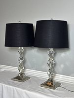 Pair of clear acrylic table lamps with black drum shades and silver-tone square bases, arranged side by side on a white surface against a grey wall.