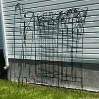 Full view of four black metal trellises and three tall black metal hooks leaning against a white siding wall on green grass.