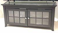 Front view of black or dark wood TV stand with two glass paneled doors and white knobs, standing on carpeted floor.