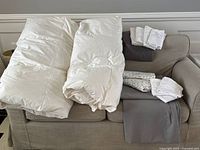 Photo of two white duvets rolled and folded on couch with folded sheets and pillowcases stacked beside them.