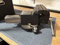 Side view of medium size black metal vice with swivel base, showing handle and jaws, on a wooden surface.