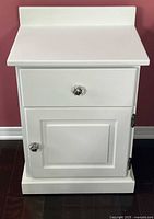 Front view of white bedside table showing drawer and closed cabinet door with glass knobs.