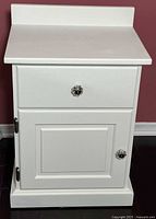 Front view of white painted wooden bedside table with drawer and single cabinet door, showing glass knobs on both drawer and door.
