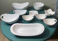 Overview of 10 white bowls and plates with side handles and varying shapes and sizes placed on a table