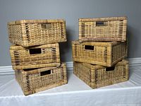 Six rectangular wicker storage baskets stacked in two piles showing front and side views, highlighting woven wicker texture and hand holes.