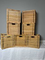 Seven wicker baskets stacked showing their fronts and handles. Natural wicker color with darker accent strips woven through.