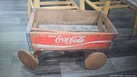 Photo of an antique Coca-Cola branded wooden wagon with Coca-Cola logo painted red on the sides on wooden wheels, sitting on a wooden floor.