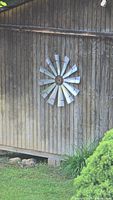 Windmill mounted on a rustic wooden exterior wall surrounded by greenery, showing the full decorative item and its outdoor placement.