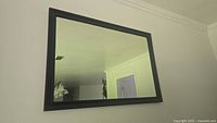 Photo showing the rectangular wall mirror mounted on a wall. The frame is black wood with smooth finish. The reflection shows part of the room.
