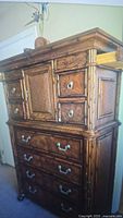 Photo front view of a traditional style warm brown wood dresser with ornate carved details, multiple drawers, central door, and decorative metal hardware. Minor wear visible.