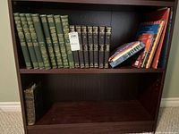 Bookshelf with multiple vintage hardcover books including two sets of volumes and several loose books stacked on right side.