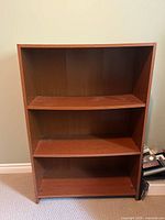 Front-facing image of a modern wooden bookshelf with three shelves, showing clear signs of wear and used condition.