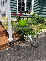 Photo taken outdoors showing front angle of three tier black metal foldable flower stand with green pot and artificial flowers on middle shelf.