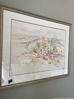 Framed watercolor landscape painting with muted earth tones and colorful foliage in front, hung on a wall.