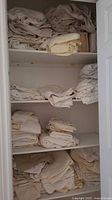 Closet shelves filled with folded king and queen size bedding in cream, beige and white colors, some loose and some in packaging.