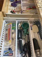 Three drawers of assorted kitchen utensils such as measuring spoons, scissors, peelers, can openers in plastic baskets.