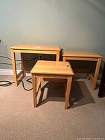 Photo showing all three wood nesting tables together on a beige carpet against a gray wall. The tables are natural wood color, rectangular with straight legs.