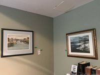 Two framed artworks on adjacent walls; left is a hand colored photograph of a harbor scene, right is a framed black and white coastal print featuring a solitary tree.