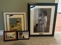 Five framed prints positioned on floor against wall. Large Moses Brooks style print of woman with child in black frame, medium silver framed girl on beach print, small comic style print and two small black and white waterfront scene prints.