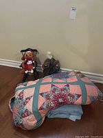 Photo showing folded LeMayne Star floral quilt on top of a blue blanket, with two Christmas figurines (Santa and dog in chair) placed behind, all on wooden floor against a beige wall.
