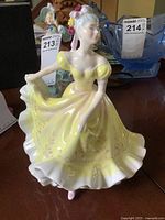 Front view of Royal Doulton Ninette HN 2379 figurine showing detailed yellow dress and figure, placed on wooden surface with other background items.