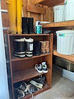 Wooden shelf with multiple pairs of boots, running shoes, ice skates, and paint cans in a garage setting.