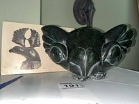Front view of dark green soapstone carving of animal head with printed provenance card in background.