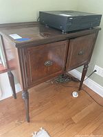 Front view of the vintage wooden cabinet showing two hinged doors with original oval metal pulls, polished wood finish, and tapered carved legs.