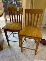 Pair of wooden pub chairs with slatted backs, natural wood finish, and foot rails shown from multiple angles on tile floor in kitchen area.