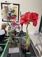 Two faux flower arrangements on display stands; one with red flowers in a glass vase, the other with white and purple flowers in a rectangular ceramic vase.